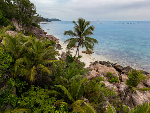 Aerial view of a tropical paradise where lush green foliage meets the turquoise sea, framed by rocky outcrops and a pristine beach, Baie Ste Anne, Praslin, Seychelles.
