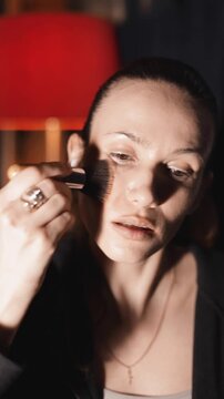 Cheerful brunette woman with a high ponytail in headphones applying contour and bronzer with a large brush on her face, natural look, smiling at night in her apartment with red lamp, vertical video