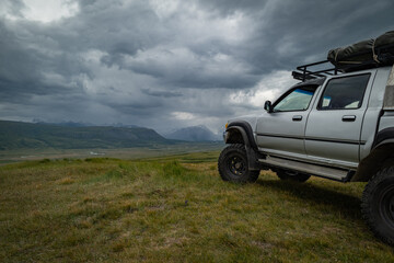 Off-road expedition vehicle standing on mountain hill under dramatic storm clouds, SUV with roof rack in vast valley with scenic mountains background, adventure travel and overlanding
