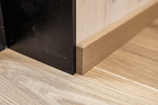 Close-up of a wooden baseboard meeting a light wood-paneled wall and floor, adjacent to a black metal frame. Natural grain contrasts with dark surface for a modern interior detail