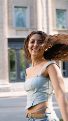 A portrait of a cheerful girl with long curly hair smiling at the camera. A close-up portrait of a friendly young woman in casual clothes against a blurred city background. Outdoors.