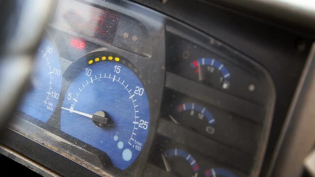Instrument panel of vehicle showing speedometer, tachometer, and fuel gauge. Red stop warning light illuminated. Surface of dashboard appears dusty. Blue and black color scheme prominent.