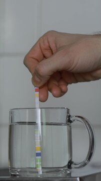 Close up view of hand dipping DIY test strip into clear water in glass mug for analysis of tap water quality, hardness, alkalinity, pH level, chlorine, nitrites, nitrates, cyanuric acid, lead in water