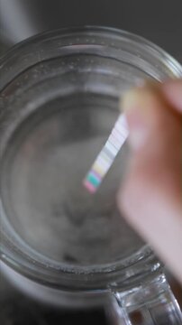 Close up, person hand holding simple test strip, immersing into clear glass of tap water. Household method determines mineral content, alkalinity, pH level, chlorine, nitrites, nitrates, hardness, cya