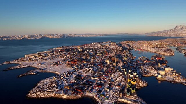 High-angle drone shot showcasing the capital city of Nuuk. Colorful houses and modern buildings spread across the snowy peninsula, surrounded by dark blue fjords and distant arctic peaks.