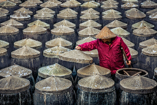 View of a person in a conical hat and red coat lifts a heavy lid from a dark vat of soy sauce / fish sauce among rows of identical vats, Xiapu, Fujian, China.