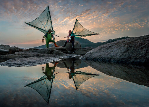View of two fishermen with triangular nets stand poised on rocky outcrops, their reflections mirrored in the still water under a dramatic sky, Xiapu, Fujian, China.