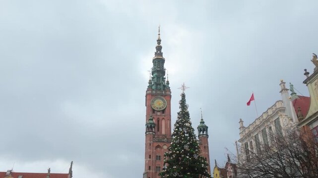 A scenic view of the historic Main Town Hall clock tower and a decorated Christmas tree in Gdansk, Poland, under an overcast winter sky.
