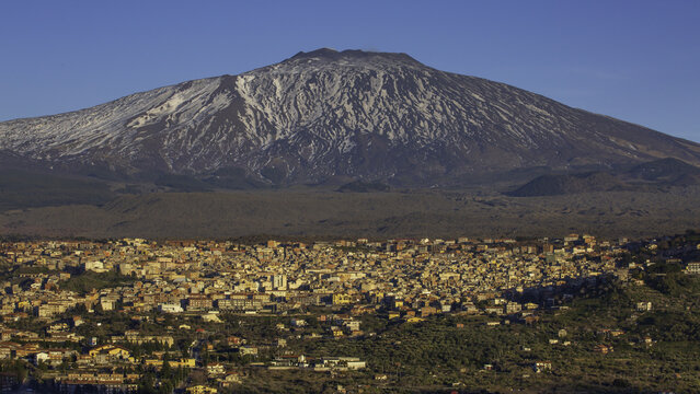 View of a sprawling town nestles beneath a majestic mountain, its peak dusted with snow, contrasting with the clear blue sky, Bronte, Sicily, Italy.