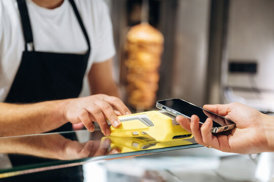 Customer making contactless payment with smartphone at food counter, cashier wearing black apron, vertical glass display case with food in background