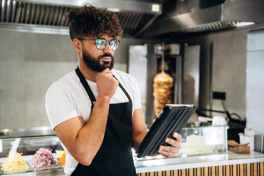 Male restaurant worker with curly hair wearing glasses checks tablet while standing in a modern kitchen with food preparation area and equipment visible in the background