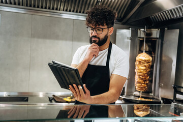 Male chef in black apron using tablet while standing in restaurant kitchen with vertical rotisserie...