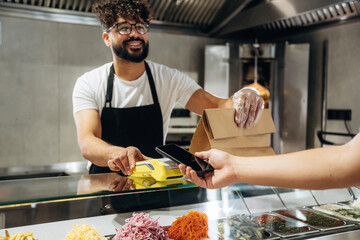 Male cashier with curly hair wearing glasses assists customer with mobile payment at a food...