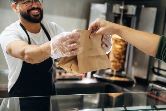 Male food vendor in black apron hands over a brown paper bag to a customer at a counter with vertical rotisserie in the background