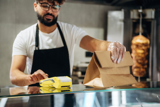 Male food service worker in black apron prepares takeout order with yellow label printer and brown paper bag in a modern restaurant setting