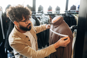 Male clothing store employee measures a brown jacket on a mannequin using a measuring tape, surrounded by various garments on display racks in a retail environment