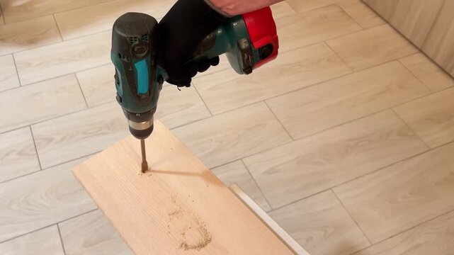 Video close-up of a gloved hand drilling a hole in a wooden plank with a cordless drill on tile floor, DIY carpentry and construction.