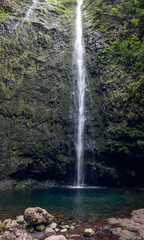 Fototapeta premium Caldeirao Verde Waterfall at trail end on route PR9, thin white plunge drops into an emerald pool beneath mossy basalt walls with ferns and soft mist (vertical panoramic photo)