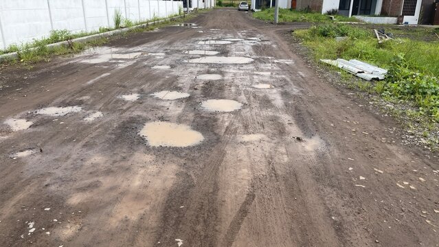 Muddy unpaved road with numerous water-filled potholes, tire tracks, and green grass on the sides, stretching towards a distant car and residential buildings.