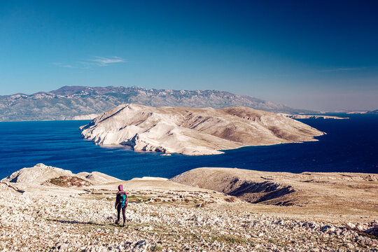 View of a lone hiker standing on a stony coastline overlooking a striking island surrounded by the deep blue sea, framed by distant mountains, Ba&Aring;&iexcl;ka, Croatia.