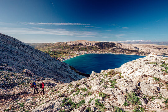 View of two hikers ascending the rocky, sun-soaked slopes towards the turquoise bay under a vast blue sky, Ba&Aring;&iexcl;ka, Primorje-Gorski Kotar County, Croatia.