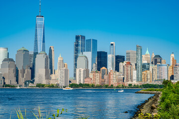 Obraz premium Lower Manhattan skyline from the shore with green foliage. Skyscrapers and One World Trade Center viewed over water with rocky shoreline and grass.