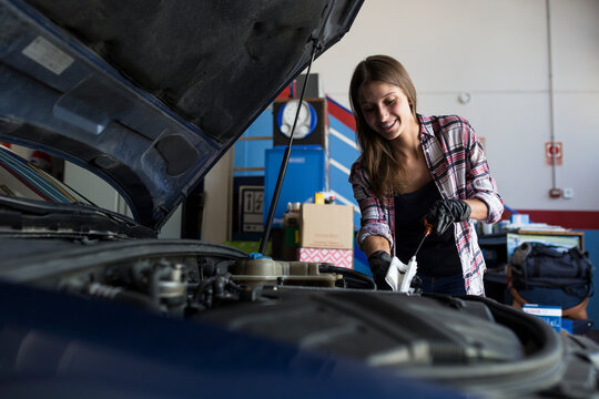 Young casual woman in shirt and gloves working with car in repair service checking engine oil level