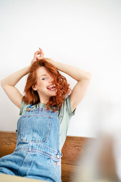 Cheerful redhead woman sitting with arms raised against white wall at home