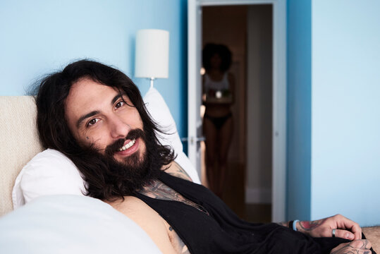 Portrait of smiling tattooed man lying n bed with girlfriend in background