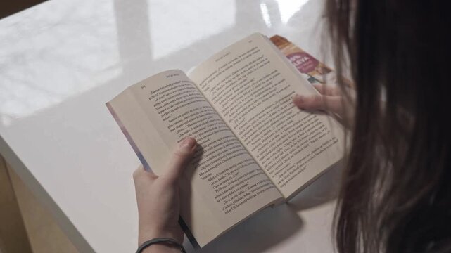 Over shoulder shot young woman reading physical book using finger swipe gesture like smartphone screen. Ideal social psychology habit, digital media addiction or literacy themes.