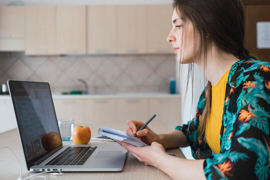 Young woman sitting at table at home using laptop and taking notes