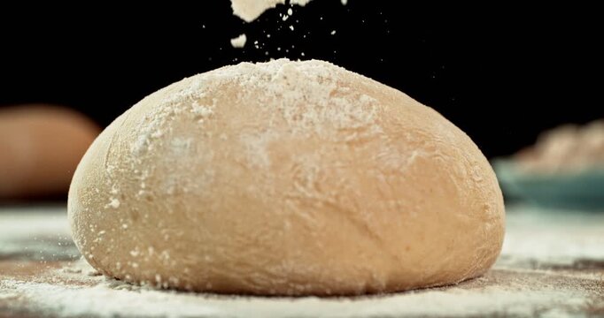 Cinematic moving shot of leavened dough isolated on black background with falling flour in super slow motion, 1000 fps. Dynamic baking concept, artisan bread preparation and texture detail.