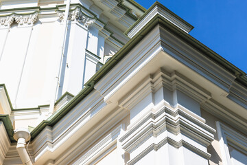 White classical building exterior with ornate cornice and layered moldings lit by sunlight against a clear blue sky, showcasing architectural detail, texture, shadow and historic design elements