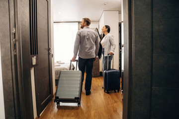 Fototapeta premium Couple entering hotel room carrying luggage, modern interior design with wooden flooring, large window providing natural light, and a cozy atmosphere visible in the background
