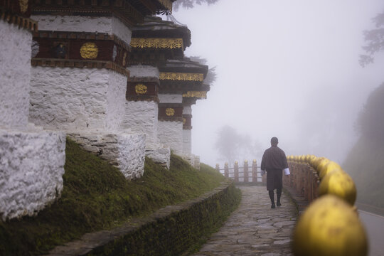 View of a solitary figure walking past ornate white chortens adorned with gold in the thick mist of Dochula, Thimphu, Bhutan.