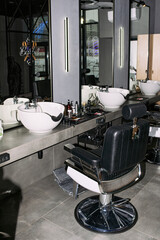 Fototapeta premium Empty barbershop station featuring modern barber chair positioned in front of large mirror with professional hair washing sink and various grooming tools, arranged on countertop