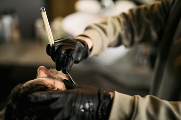 Caucasian young adult man receiving professional shave from gloved barber using straight razor in barbershop, barber carefully shaping facial hair while client reclines in chair