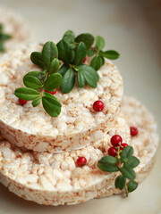 Close-up of lingonberry (Vaccinium vitis-idaea) sprigs with red berries and green leaves arranged on stacked rice cakes. Natural plant decoration and healthy snack concept in soft light.
