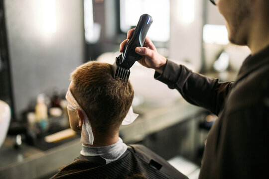 Caucasian young adult man sitting in barbershop chair receiving haircut from Caucasian young adult male barber using electric hair clipper, both focused on grooming process