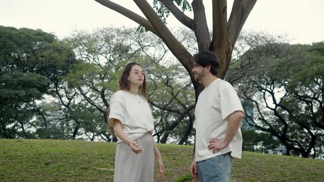 Young couple gives high five slapping palms together joyfully beside massive tree trunk on sloping grassy hill in verdant park amid surrounding leafy branches and open lawn expanse.