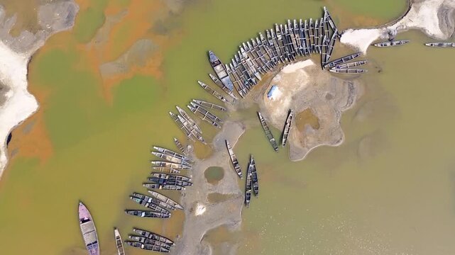 Aerial view of numerous boats clustered along the riverbank, creating a textural contrast with the muddy shore and the green-brown water, Sunamganj, Sylhet Division, Bangladesh.