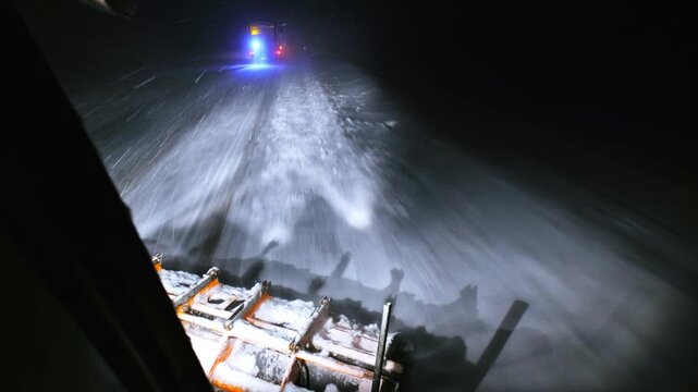 POV from a snowplow clearing a highway at night during a severe winter storm. Bright headlights, blowing snow and low visibility emphasize hazardous weather and ongoing road maintenance work.