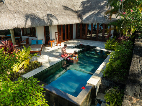 Aerial view of a couple enjoying a floating meal in a pool surrounded by lush greenery and a thatched-roof villa, Prince Maurice, Flacq District, Mauritius.