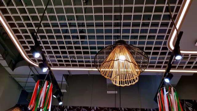 Warm woven pendant lights and track lighting in a modern restaurant interior. Festive tricolor ribbons hang from a grid ceiling above a patterned wall decor.