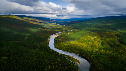 Fototapeta premium A winding river carves its path through a lush green valley under a cloudy sky.