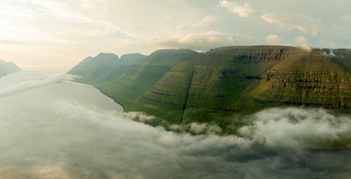 Aerial view of majestic green mountains rise dramatically from the tranquil waters, embraced by ethereal clouds, creating a serene yet powerful landscape, Klaksvik, Northern Isles, Faroe Islands.