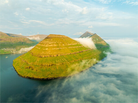 Aerial view of the majestic, tiered island rising from the sea, shrouded in ethereal mist, its vibrant green slopes contrasting with the deep blue waters, Klaksvik, Faroe Islands.