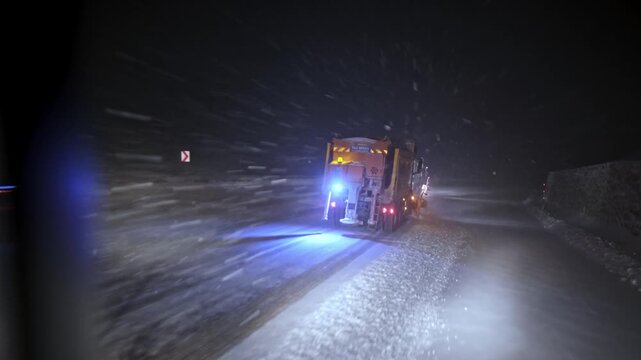 View from inside a vehicle following a snowplow clearing a snow-covered road at night. Blowing snow, flashing warning lights and low visibility show dangerous winter driving and road maintenance opera