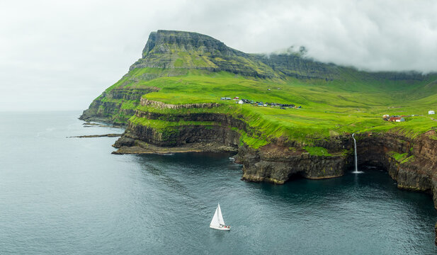 Aerial view of a solitary sailboat glides past the towering cliffs where the Mulafossur waterfall plunges into the sea, Gasadalur, Vagar, Faroe Islands.