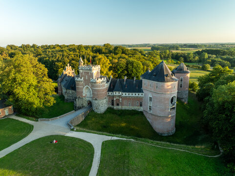 Aerial view of Gaasbeek Castle's red brick walls rise amidst lush green forests, a bridge arching towards its grand entrance, Lennik, Vlaanderen, Belgium.